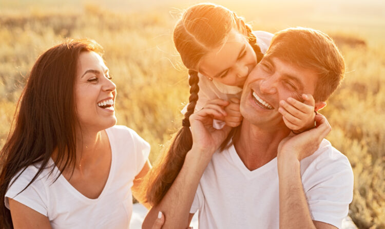 Daughter hugging and kissing cheerful father in cheek near laughing mother while spending day of happy childhood in field