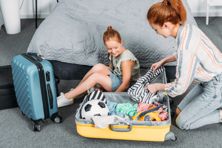 happy mother trying to pack clothes into suitcase while daughter sitting in it